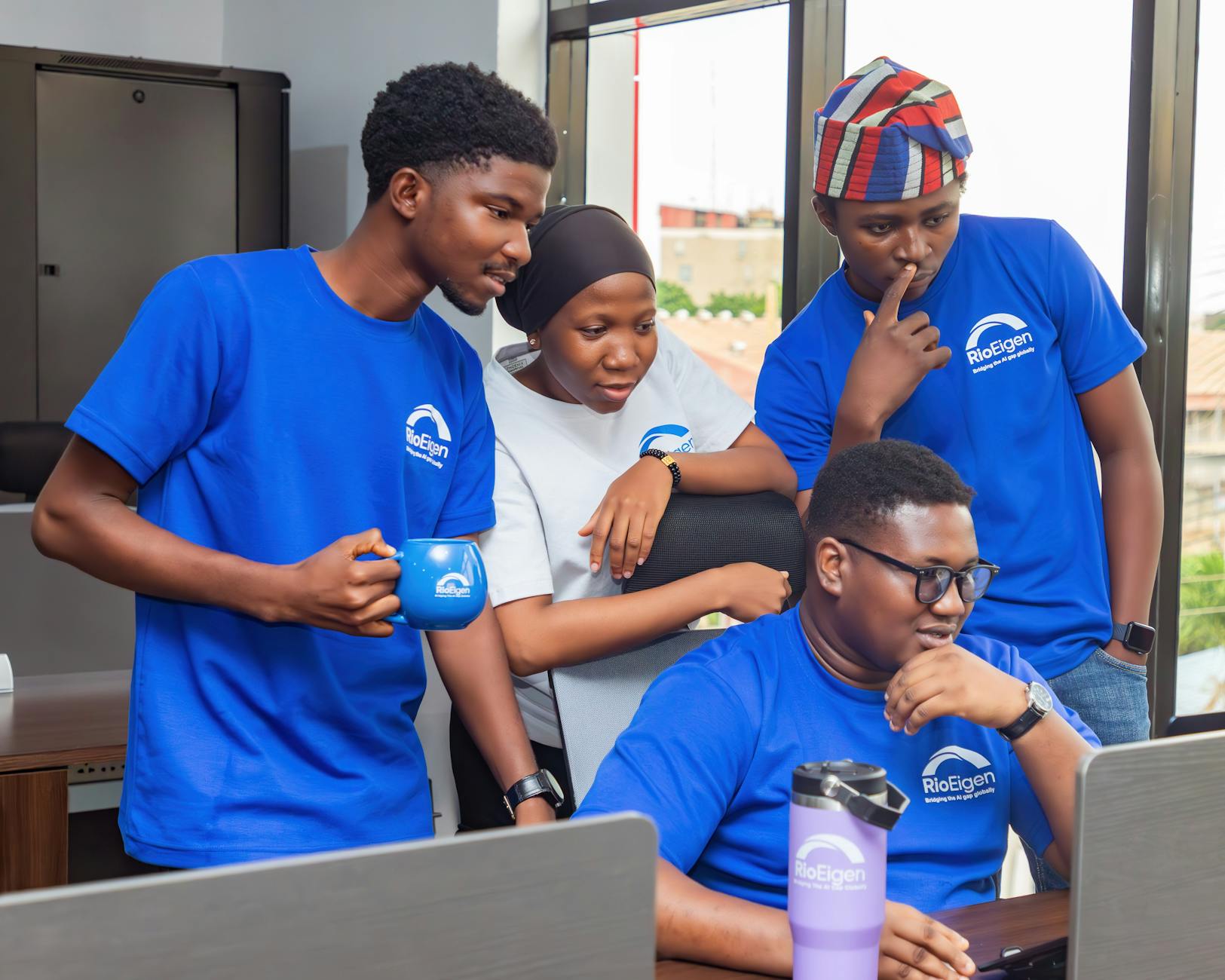 A Diverse Team Of Adults Collaborating Around A Computer In A Modern Office Setting.