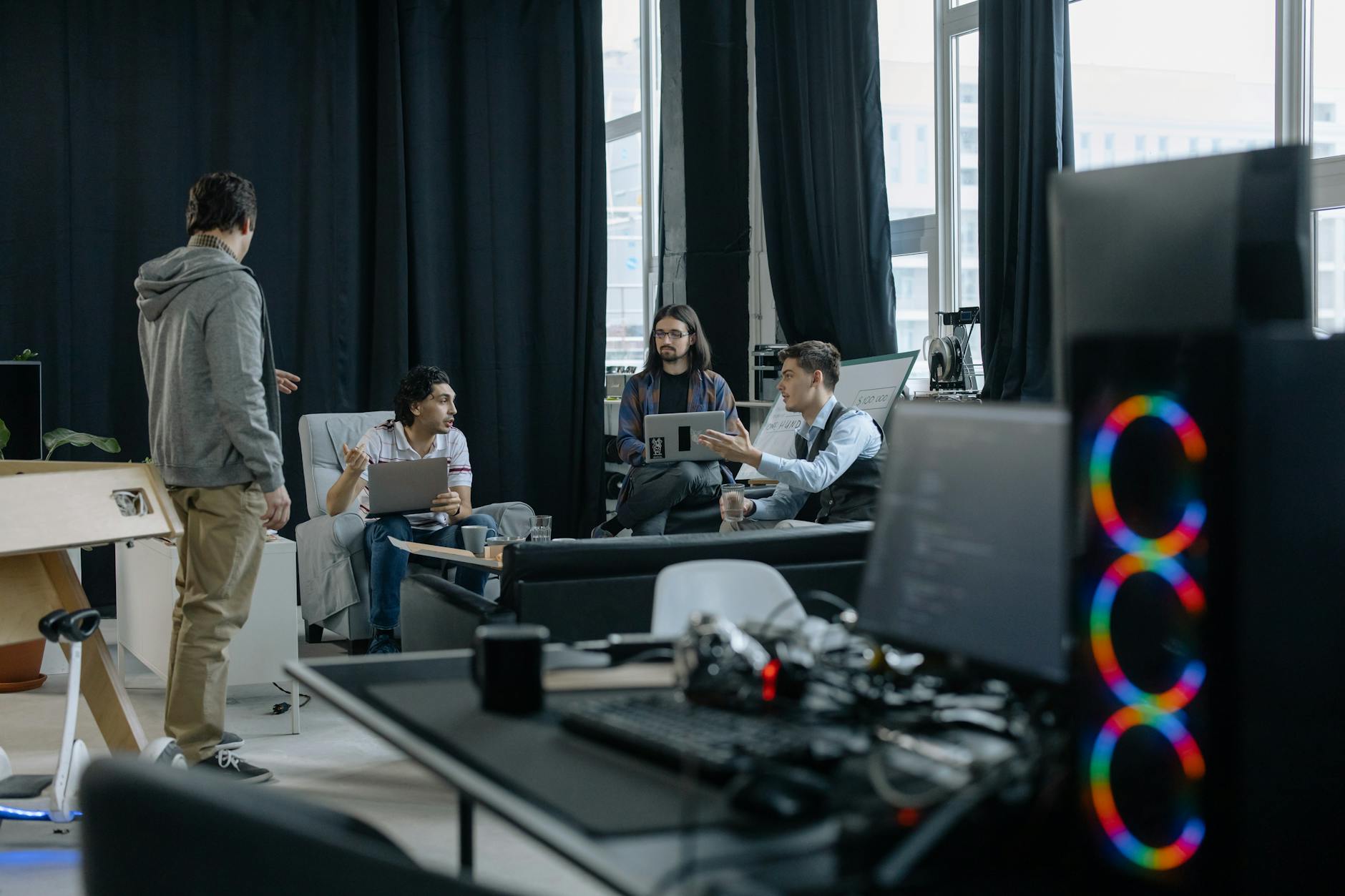 A Group Of Professionals Engaged In A Casual Meeting At A Modern Office, Discussing Projects On Laptops And Tablets.