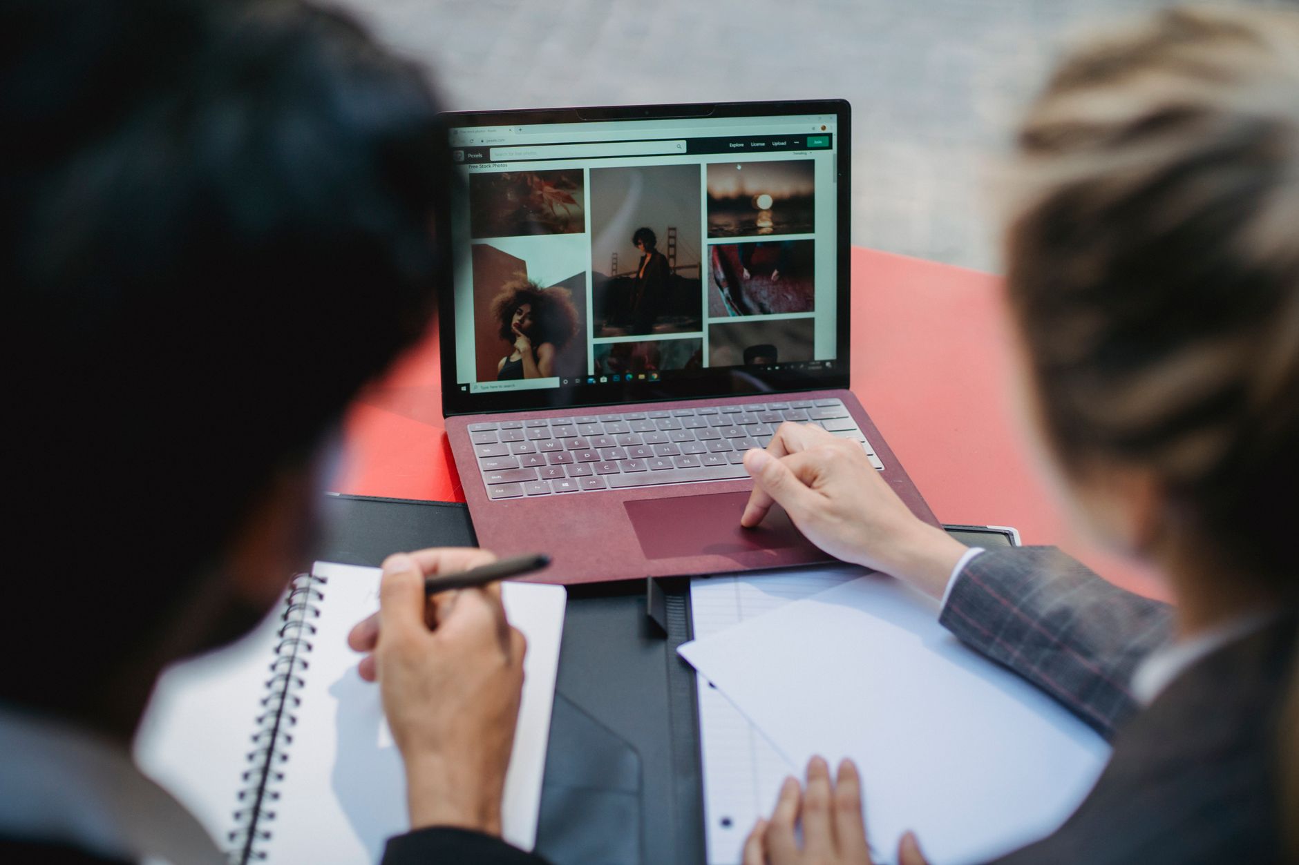 Two Professionals Working On A Laptop At A Table, Discussing Content And Taking Notes.