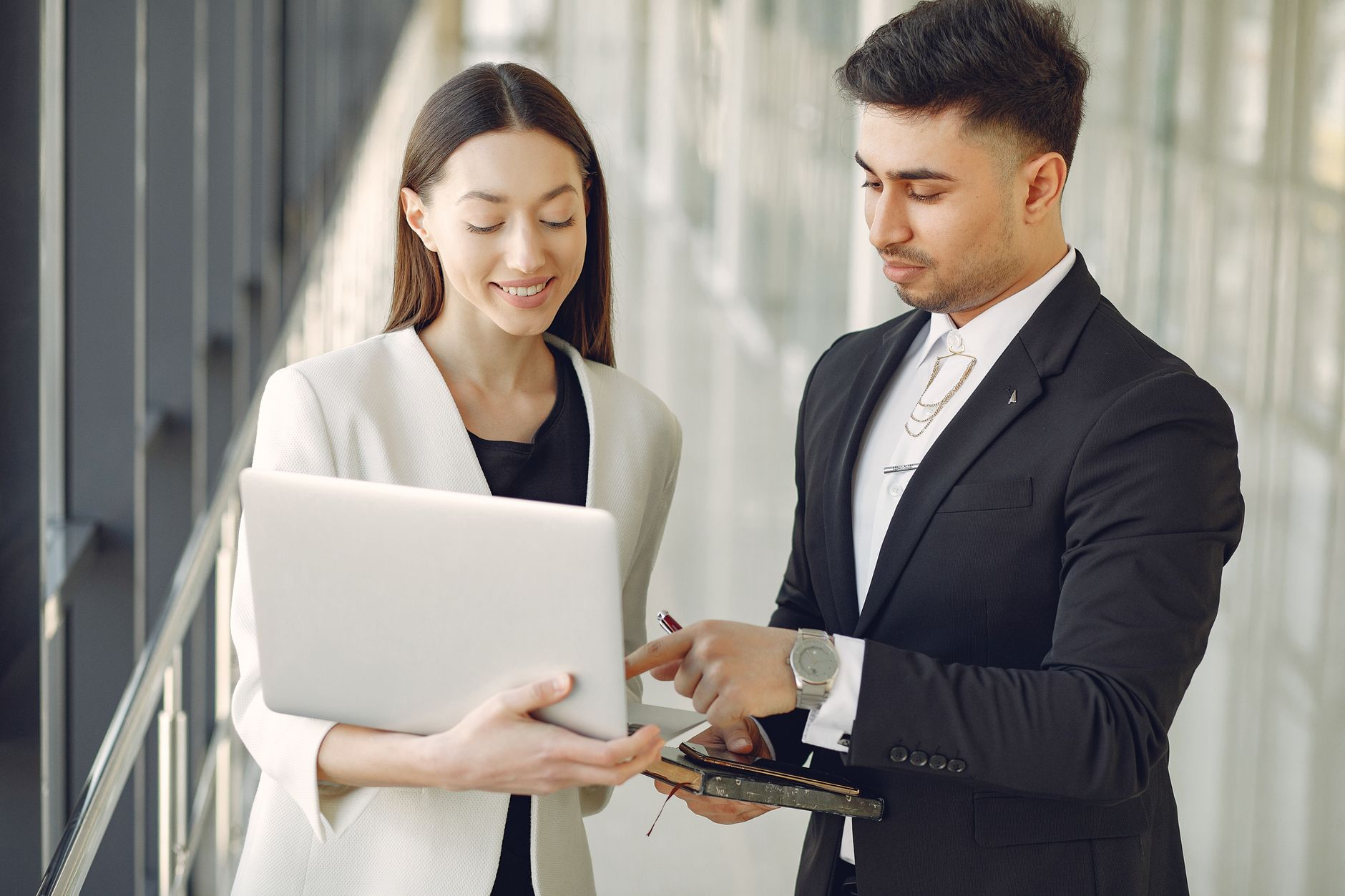 Concentrated Diverse Male And Female Employees In Formal Clothes Standing In Modern Office Corridor And Browsing Laptop While Discussing Business Project Together