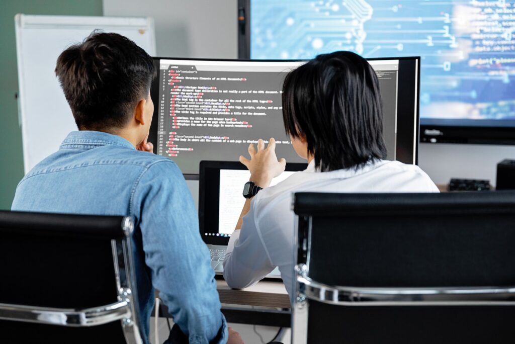 Two Developers Working Together In An Office, Discussing Code On A Screen. Technology Teamwork Setting.